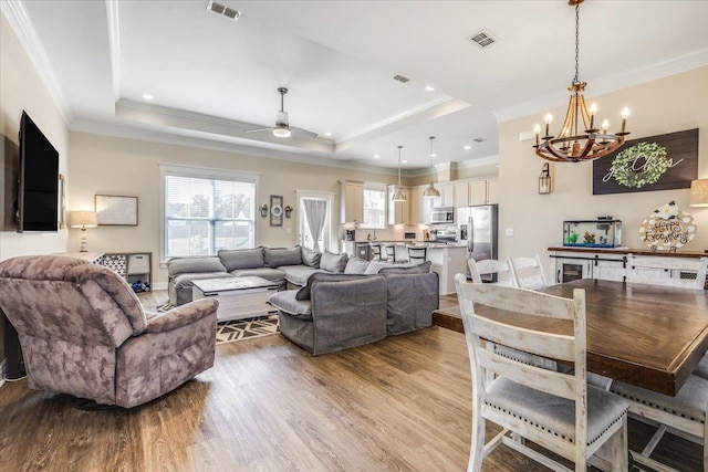 living room with light wood-type flooring, crown molding, ceiling fan with notable chandelier, and a raised ceiling