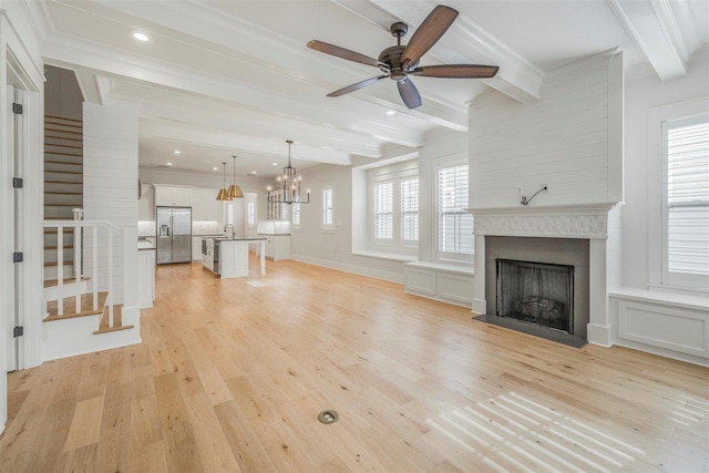 unfurnished living room with a fireplace, stairway, light wood-type flooring, beamed ceiling, and ceiling fan with notable chandelier