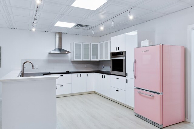 kitchen featuring white fridge, stainless steel oven, wall chimney range hood, white cabinetry, and light wood-type flooring