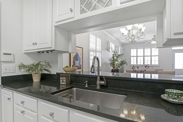 kitchen featuring dark stone counters, ornamental molding, ceiling fan with notable chandelier, white cabinets, and a sink