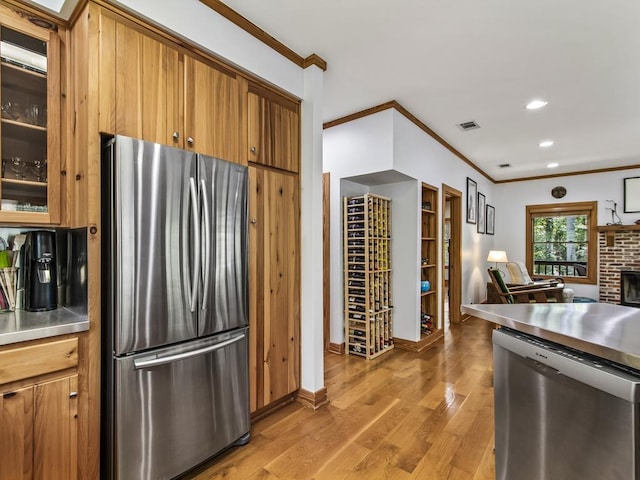 kitchen with crown molding, stainless steel counters, appliances with stainless steel finishes, a fireplace, and light hardwood / wood-style floors