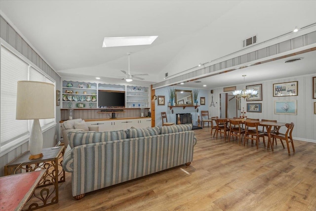 living room with built in shelves, ceiling fan with notable chandelier, light hardwood / wood-style floors, and vaulted ceiling with skylight