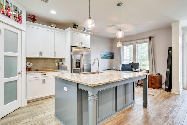 kitchen with sink, stainless steel fridge, white cabinetry, light stone countertops, and a center island with sink