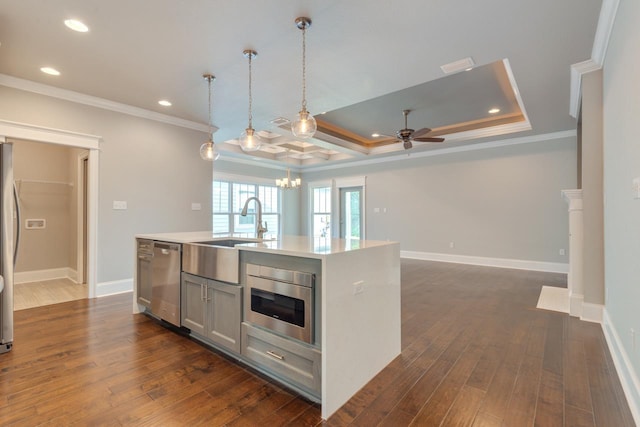 kitchen with appliances with stainless steel finishes, a raised ceiling, a kitchen island with sink, crown molding, and pendant lighting