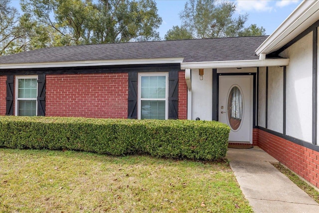 view of exterior entry featuring brick siding, stucco siding, a lawn, and roof with shingles