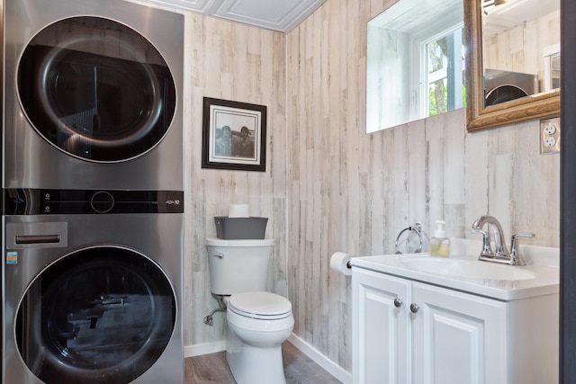 bathroom featuring toilet, stacked washer and clothes dryer, hardwood / wood-style floors, wooden walls, and vanity