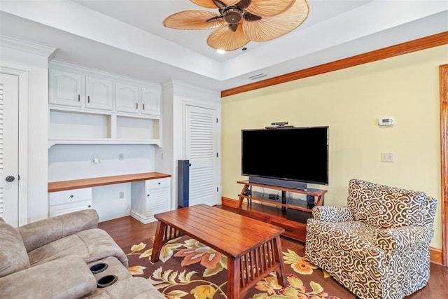 living room with crown molding, ceiling fan, a tray ceiling, built in desk, and dark hardwood / wood-style flooring