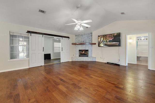 unfurnished living room featuring hardwood / wood-style floors, vaulted ceiling, a stone fireplace, ceiling fan, and a barn door