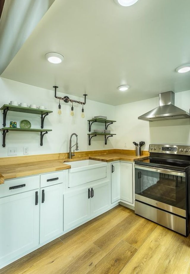 kitchen featuring butcher block counters, wall chimney range hood, stainless steel range with electric cooktop, light hardwood / wood-style floors, and white cabinets