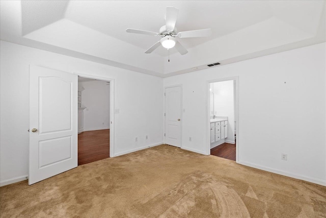 unfurnished bedroom featuring ceiling fan, ensuite bath, carpet flooring, and a tray ceiling