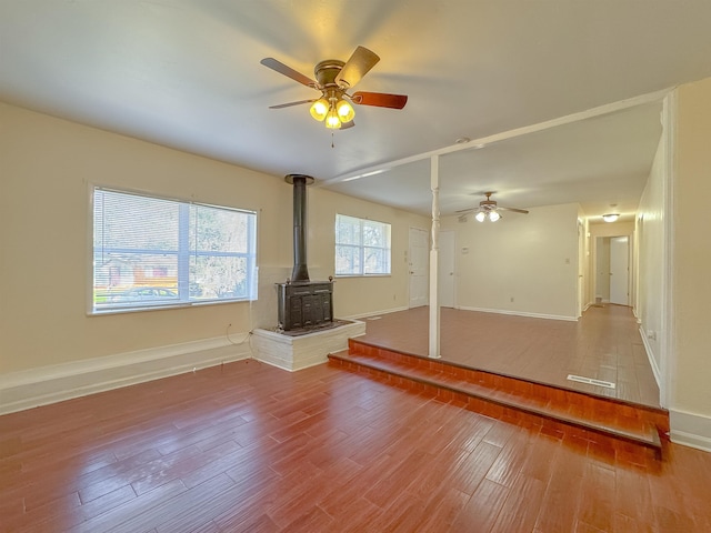 unfurnished living room with baseboards, ceiling fan, wood finished floors, and a wood stove