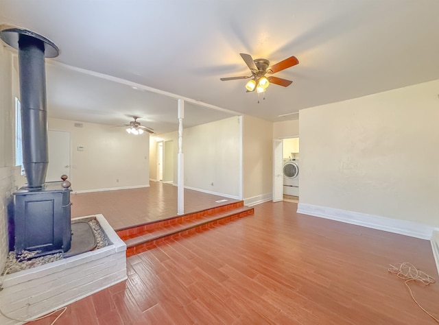 unfurnished living room with baseboards, washer / dryer, a wood stove, wood finished floors, and a ceiling fan