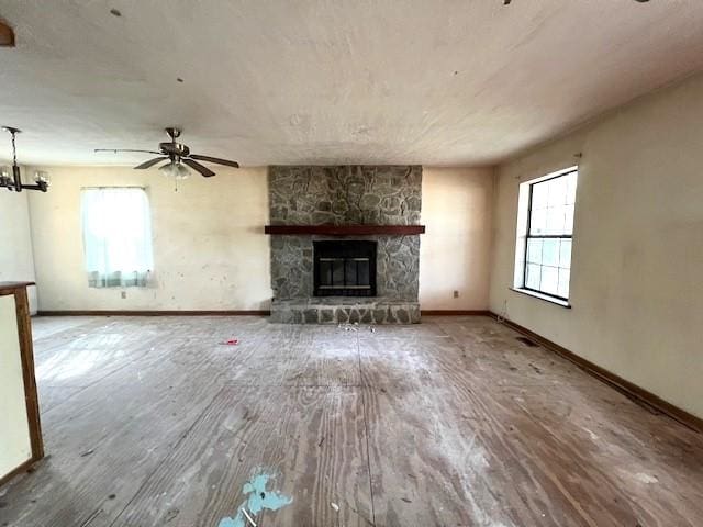 unfurnished living room with ceiling fan with notable chandelier, a stone fireplace, baseboards, and wood finished floors