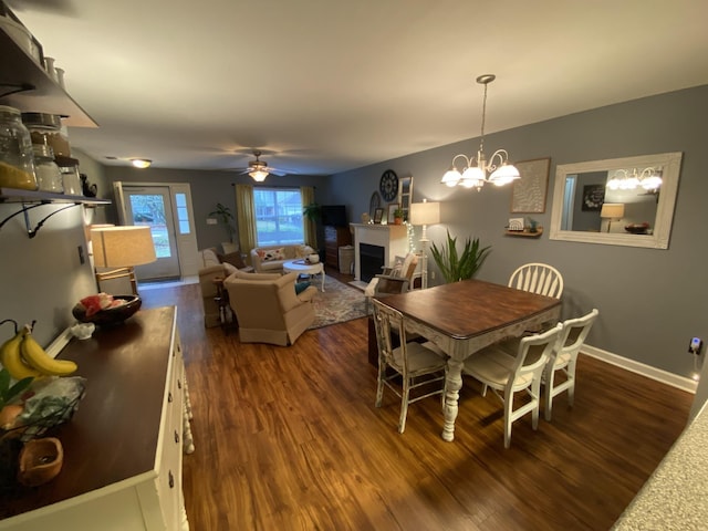 dining area featuring dark wood-style floors, ceiling fan with notable chandelier, a fireplace, and baseboards