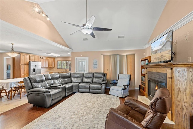 living area with visible vents, high vaulted ceiling, a warm lit fireplace, light wood finished floors, and ceiling fan