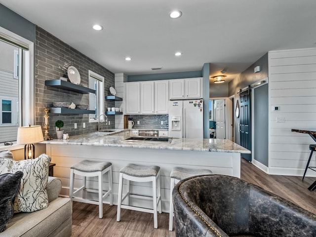 kitchen featuring white fridge with ice dispenser, open shelves, a sink, a peninsula, and light stone counters