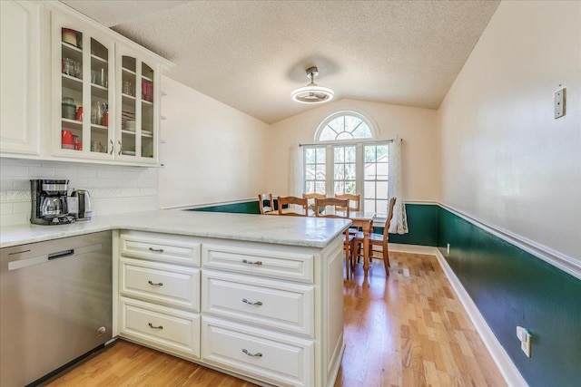 kitchen with light countertops, stainless steel dishwasher, glass insert cabinets, vaulted ceiling, and a peninsula