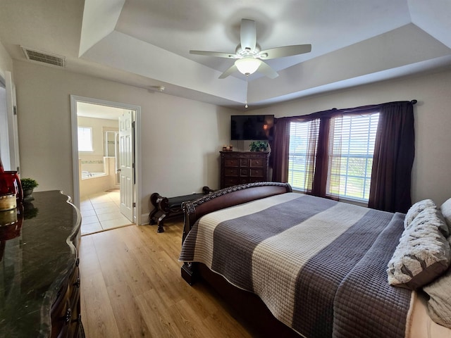 bedroom with ensuite bathroom, light wood-style flooring, visible vents, a ceiling fan, and a tray ceiling