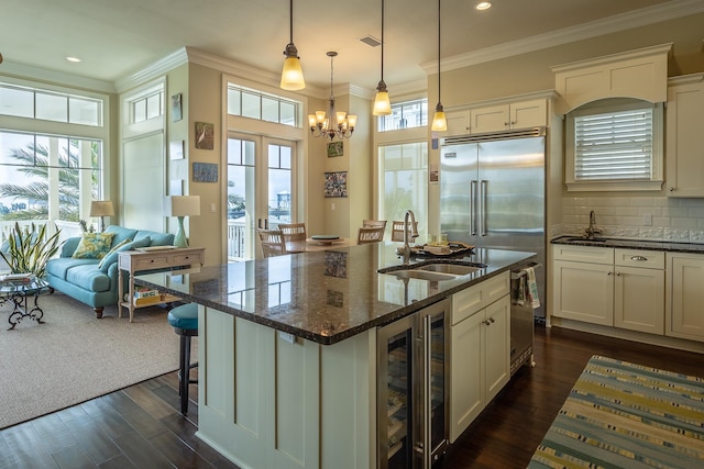 kitchen with an island with sink, sink, beverage cooler, dark stone counters, and crown molding