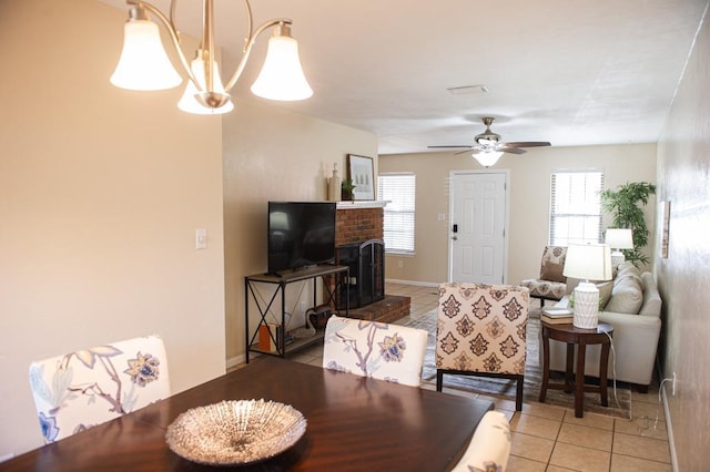 dining space with ceiling fan with notable chandelier, a healthy amount of sunlight, light tile patterned floors, and a brick fireplace