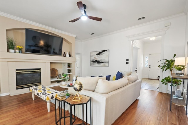living area featuring a ceiling fan, visible vents, crown molding, a tiled fireplace, and wood finished floors