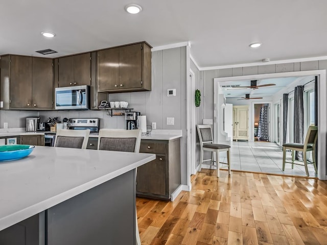 kitchen with ceiling fan, stainless steel appliances, dark brown cabinetry, ornamental molding, and light hardwood / wood-style floors