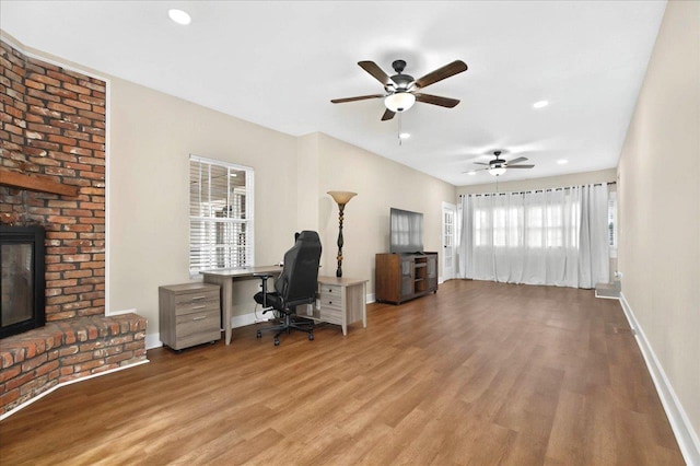 office space with baseboards, a brick fireplace, wood finished floors, and a ceiling fan