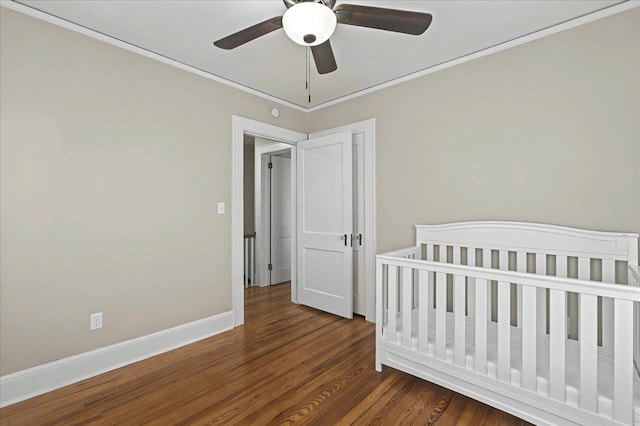 bedroom featuring a nursery area, dark wood-style flooring, crown molding, baseboards, and ceiling fan