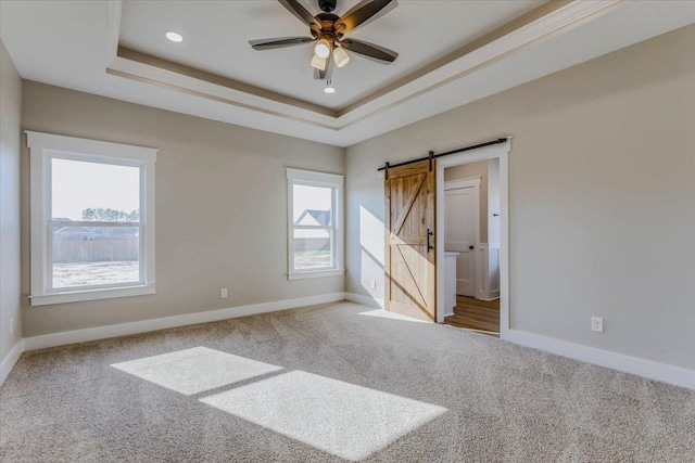 unfurnished bedroom with a barn door, ceiling fan, a tray ceiling, and carpet