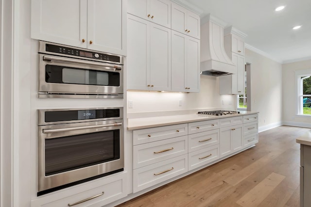 kitchen featuring crown molding, appliances with stainless steel finishes, white cabinetry, premium range hood, and light hardwood / wood-style floors