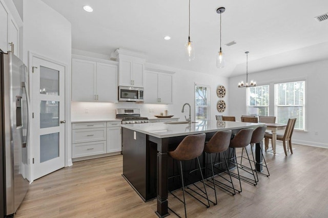 kitchen featuring stainless steel appliances, light wood-type flooring, decorative light fixtures, an island with sink, and white cabinets