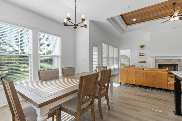 dining space with a stone fireplace, ceiling fan with notable chandelier, light hardwood / wood-style floors, wooden ceiling, and a raised ceiling