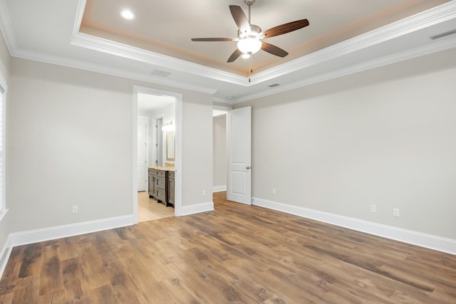 unfurnished bedroom with ensuite bath, ornamental molding, ceiling fan, a tray ceiling, and light wood-type flooring