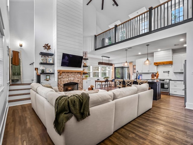 living area with ceiling fan, dark wood-style flooring, a towering ceiling, a brick fireplace, and recessed lighting