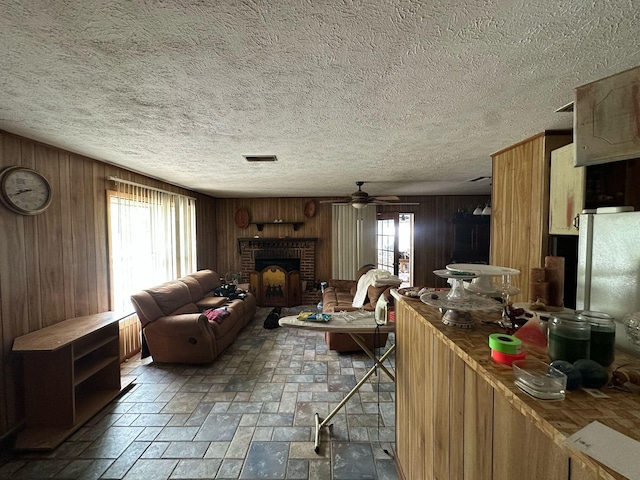 living room featuring a textured ceiling, wood walls, visible vents, ceiling fan, and a brick fireplace