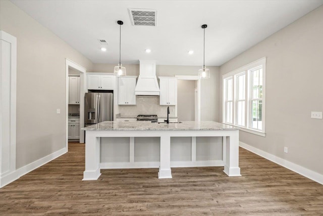 kitchen with custom exhaust hood, white cabinetry, stainless steel appliances, and a kitchen island with sink