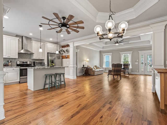 interior space with ornate columns, appliances with stainless steel finishes, white cabinetry, a raised ceiling, and wall chimney range hood