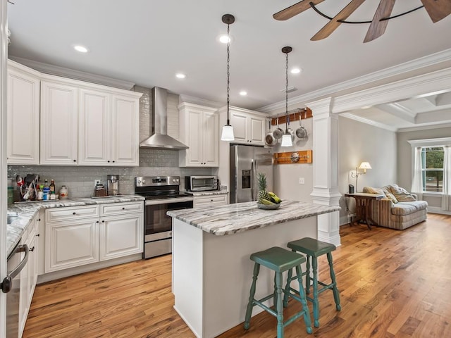 kitchen with appliances with stainless steel finishes, a kitchen island, wall chimney range hood, decorative columns, and white cabinets