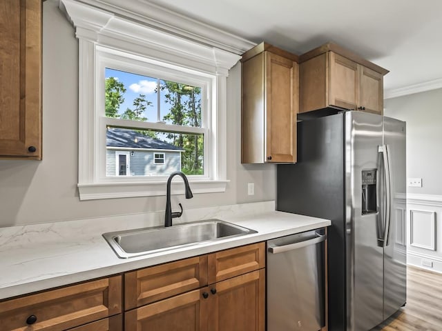 kitchen with appliances with stainless steel finishes, sink, light stone counters, crown molding, and light hardwood / wood-style flooring