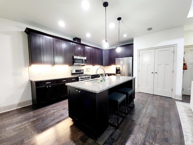 kitchen with appliances with stainless steel finishes, dark hardwood / wood-style flooring, light stone counters, a kitchen island with sink, and sink