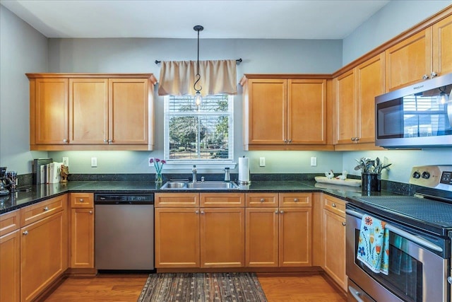 kitchen with appliances with stainless steel finishes, sink, decorative light fixtures, dark stone counters, and light hardwood / wood-style floors