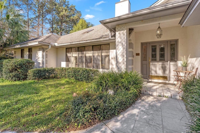 view of exterior entry featuring a shingled roof, a lawn, a chimney, and stucco siding