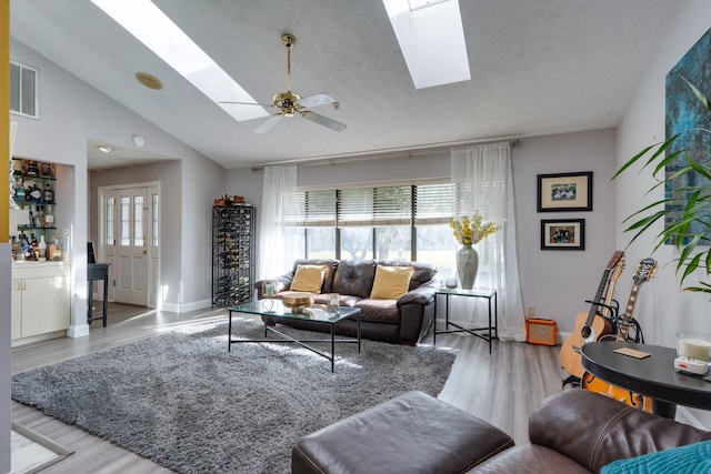 living room featuring a ceiling fan, wood finished floors, visible vents, vaulted ceiling with skylight, and a textured ceiling