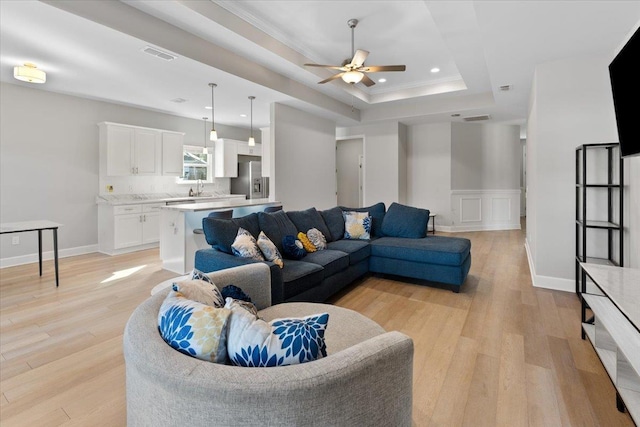 living room with sink, crown molding, light wood-type flooring, a raised ceiling, and ceiling fan