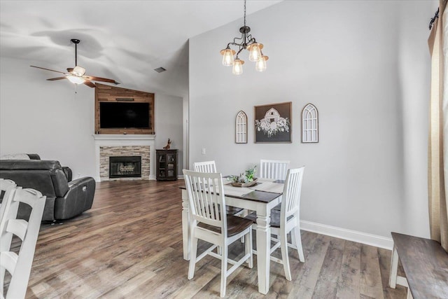 dining area featuring ceiling fan with notable chandelier, wood-type flooring, a stone fireplace, and vaulted ceiling