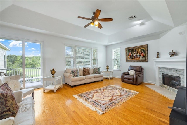 living room featuring light wood-type flooring, a tray ceiling, a stone fireplace, and ceiling fan