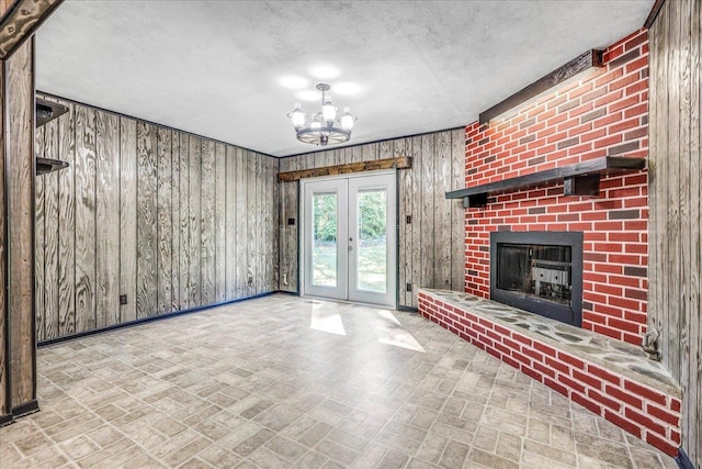 unfurnished living room with wooden walls, a textured ceiling, french doors, a brick fireplace, and a chandelier