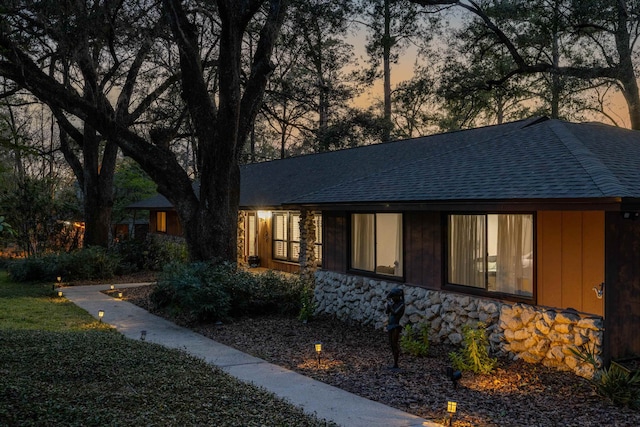 property exterior at dusk featuring stone siding and roof with shingles