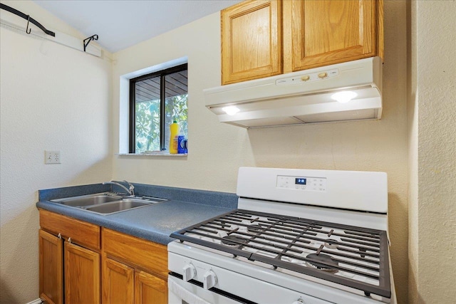 kitchen with dark countertops, brown cabinets, white gas stove, under cabinet range hood, and a sink