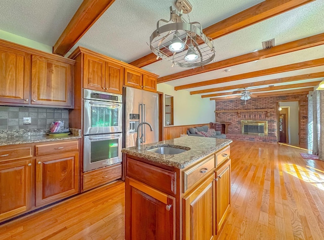 kitchen featuring a kitchen island with sink, sink, a brick fireplace, appliances with stainless steel finishes, and beamed ceiling